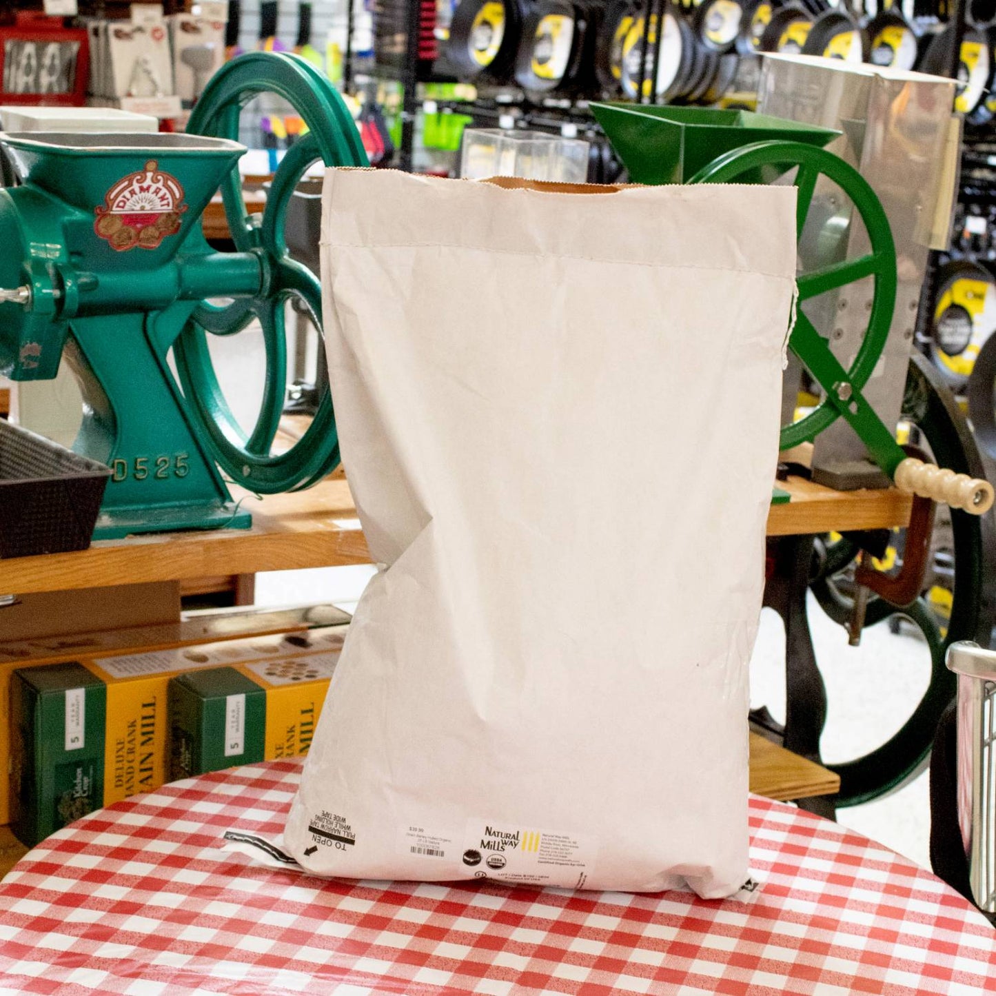 Large plain white paper bag filled with grain, set on a red and white checkered tablecloth with vintage green hand-crank grain mills and kitchen tools in the background, highlighting pantry and grain mill collections.