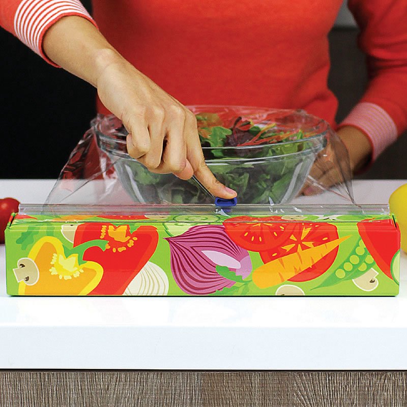Hand sliding the blue cutter across a reusable dispenser of plastic wrap with colorful vegetable illustrations, covering a glass bowl of fresh greens, Kitchen & Food and Storage collections.