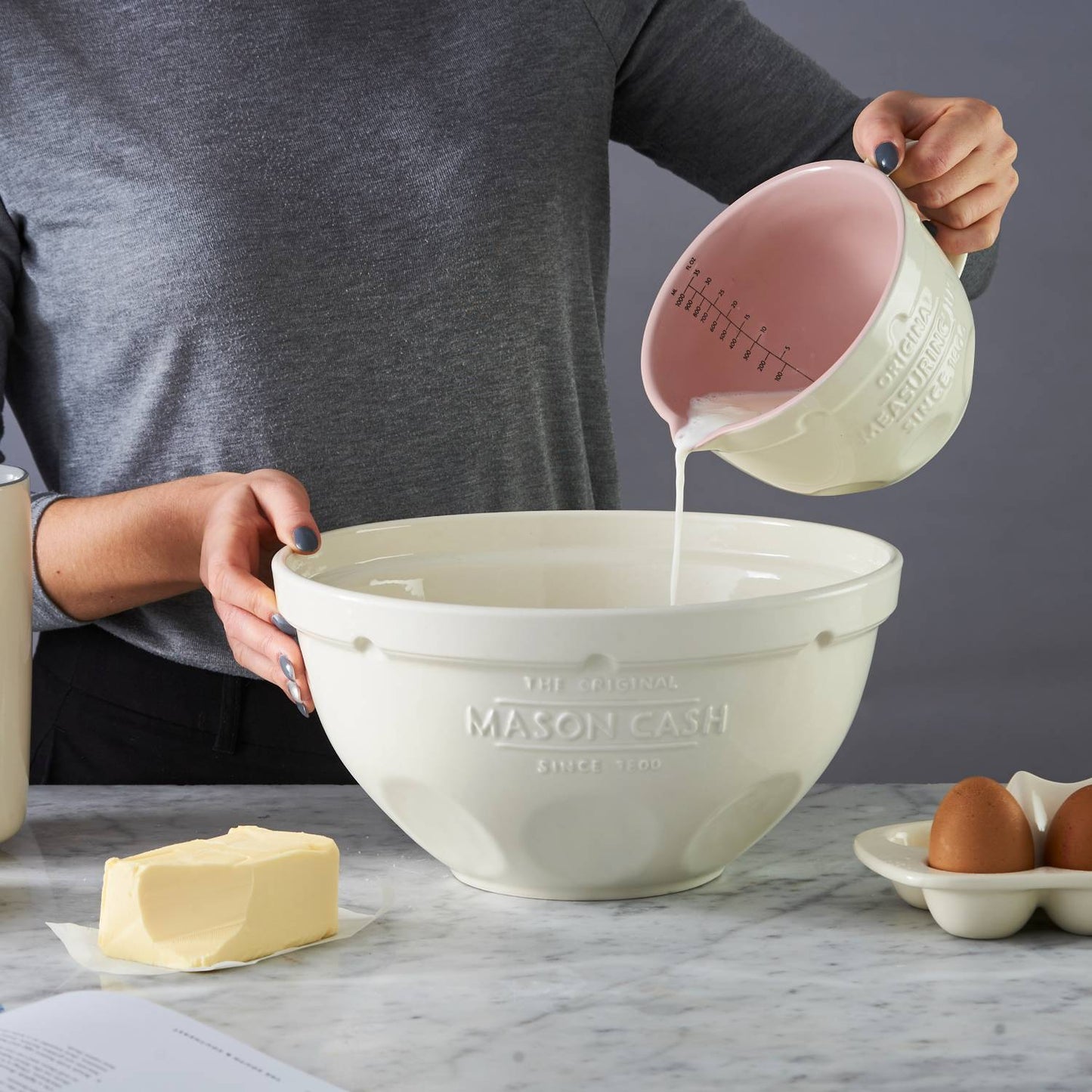 Cream-colored measuring jug with pink interior gently pouring liquid into matching mixing bowl on marble countertop, surrounded by butter and eggs, featured in Kitchen & Food, Cookware, Baking Supplies, Food Prep, Measuring, Best Sellers, Promo Eligible collections.