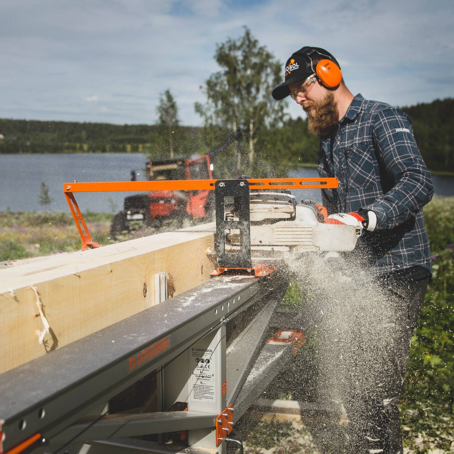 Man wearing safety glasses and earmuffs operating a LOGOSOL chainsawmill with an orange bar nose top steering accessory, cutting wood outdoors near a lake, in Farm, Garden & Tool and Wood Cutting and Hauling collections.