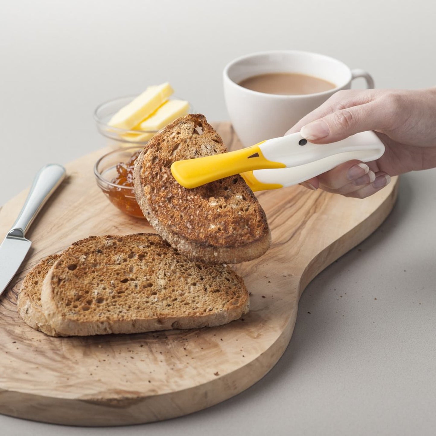 Handheld duck-shaped toast tongs with a heat-resistant yellow silicone tip gripping a toasted bread slice on a wooden serving board, shown with tea and butter in Kitchen & Food collections.