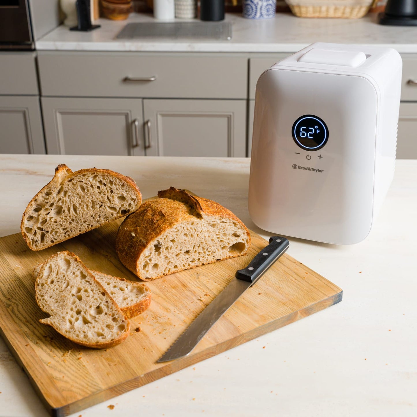 Compact white temperature-controlled device displaying 62°F on a digital screen, placed next to freshly sliced artisan bread and a serrated knife on a wooden cutting board, ideal for Kitchen & Food, Baking Supplies, and Kitchen Gadgets collections.