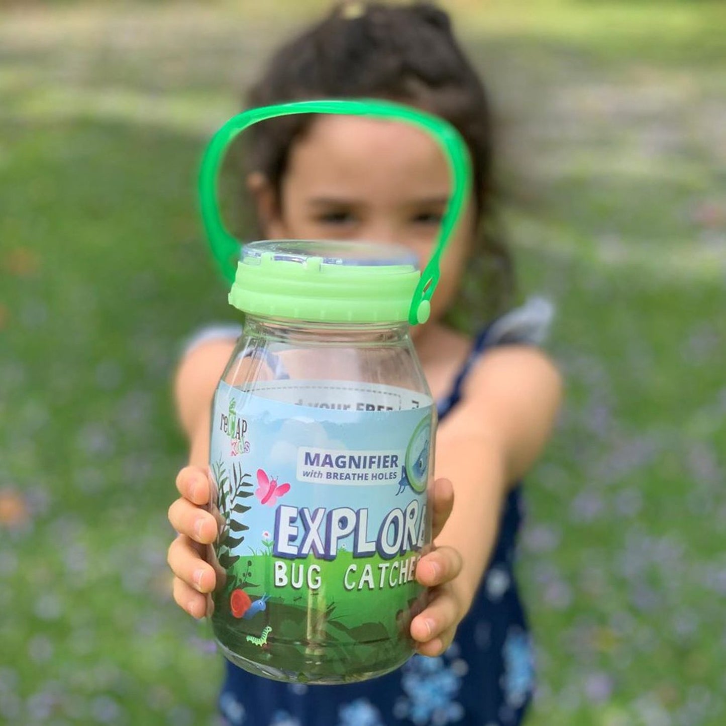 Child outdoors holding a handcrafted clear plastic bug catcher with a green glow-in-the-dark magnifying lid and breathable cap, encouraging outdoor play and exploration in USA Made toys from Lehman’s collection.