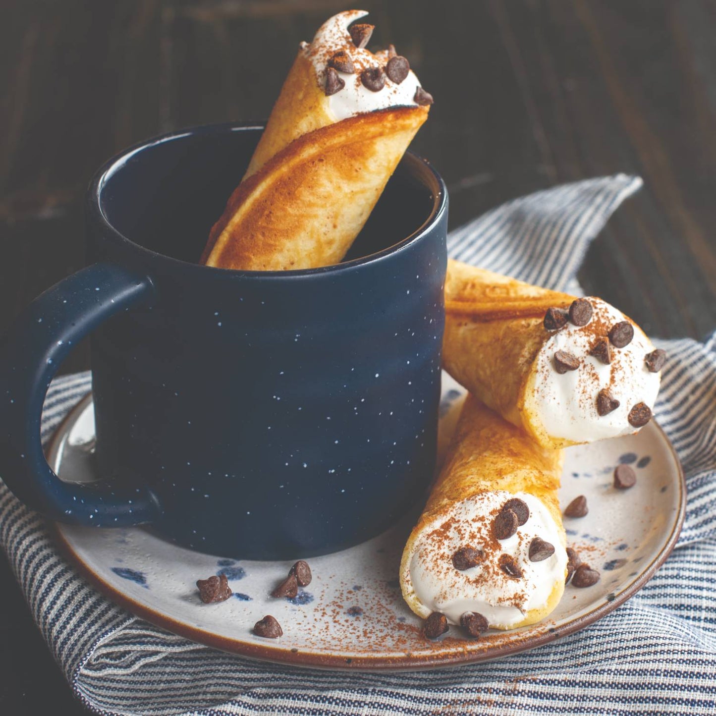 Three handcrafted krumkake cookies filled with whipped cream and chocolate chips, served on a speckled plate next to a dark blue ceramic mug, styled for Kitchen & Food and Baking Supplies collections.