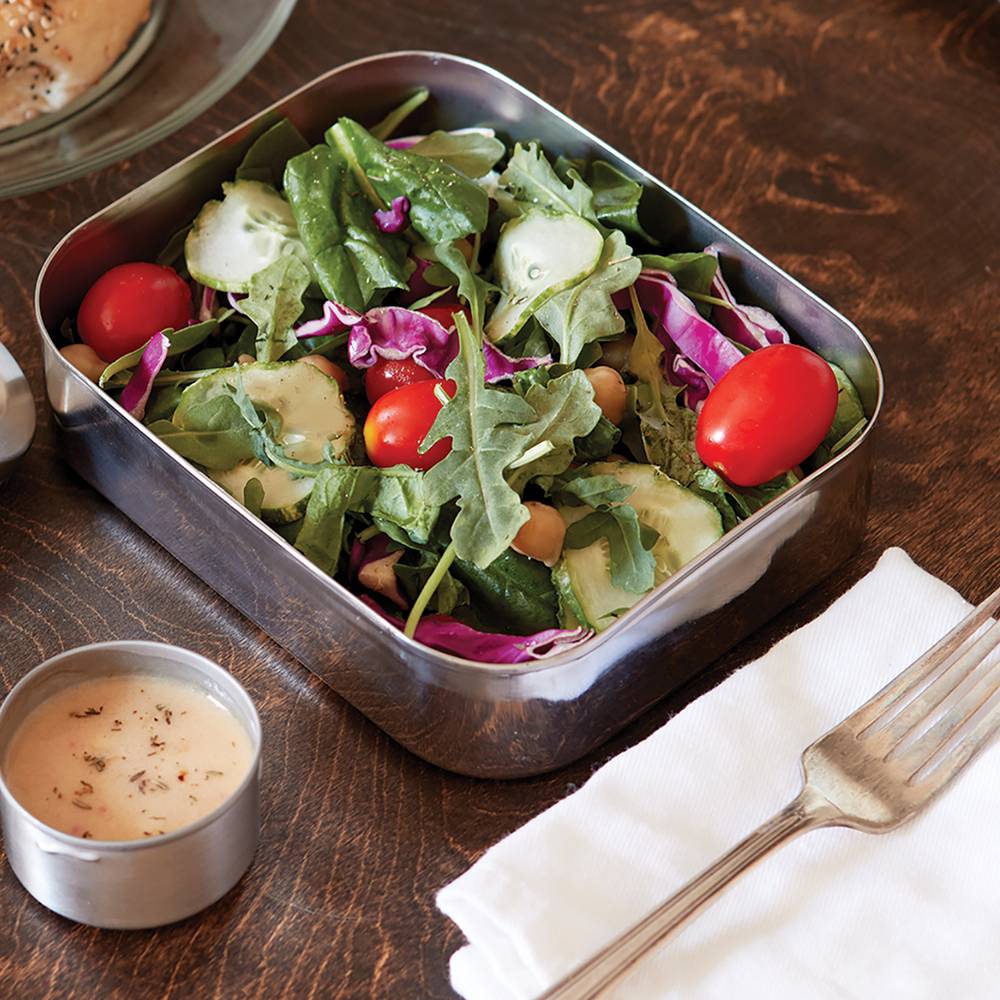 Open stainless steel container filled with fresh mixed salad greens, cherry tomatoes, cucumber slices, and red cabbage, accompanied by a small dipping sauce cup, fork, and napkin on a wooden table, featured in Kitchen & Food and Storage and Serving collections.