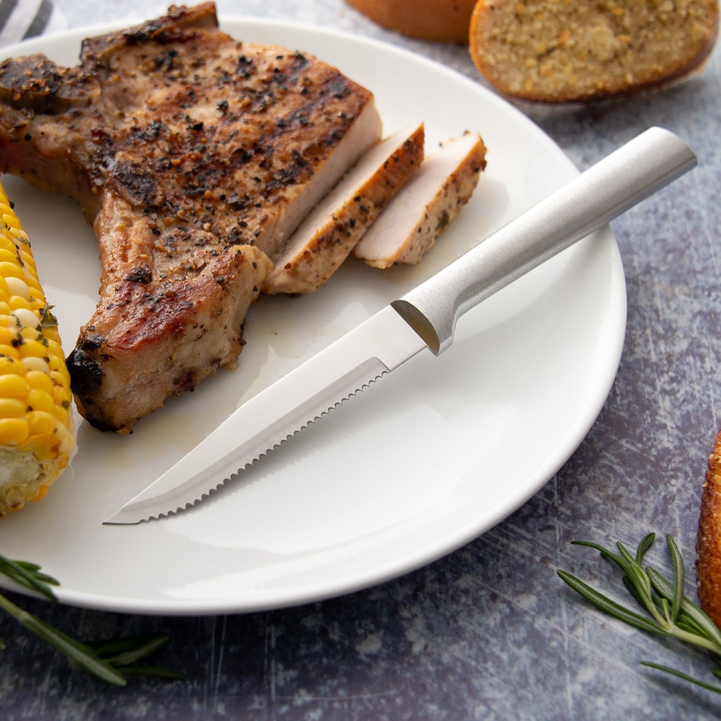Handcrafted Rada serrated steak knife with high carbon surgical stainless steel blade and solid aluminum handle slicing a grilled pork chop, served with corn on the cob and bread, ideal for Kitchen & Food, Farm, Garden & Tool, USA Made, and Best Sellers collections.