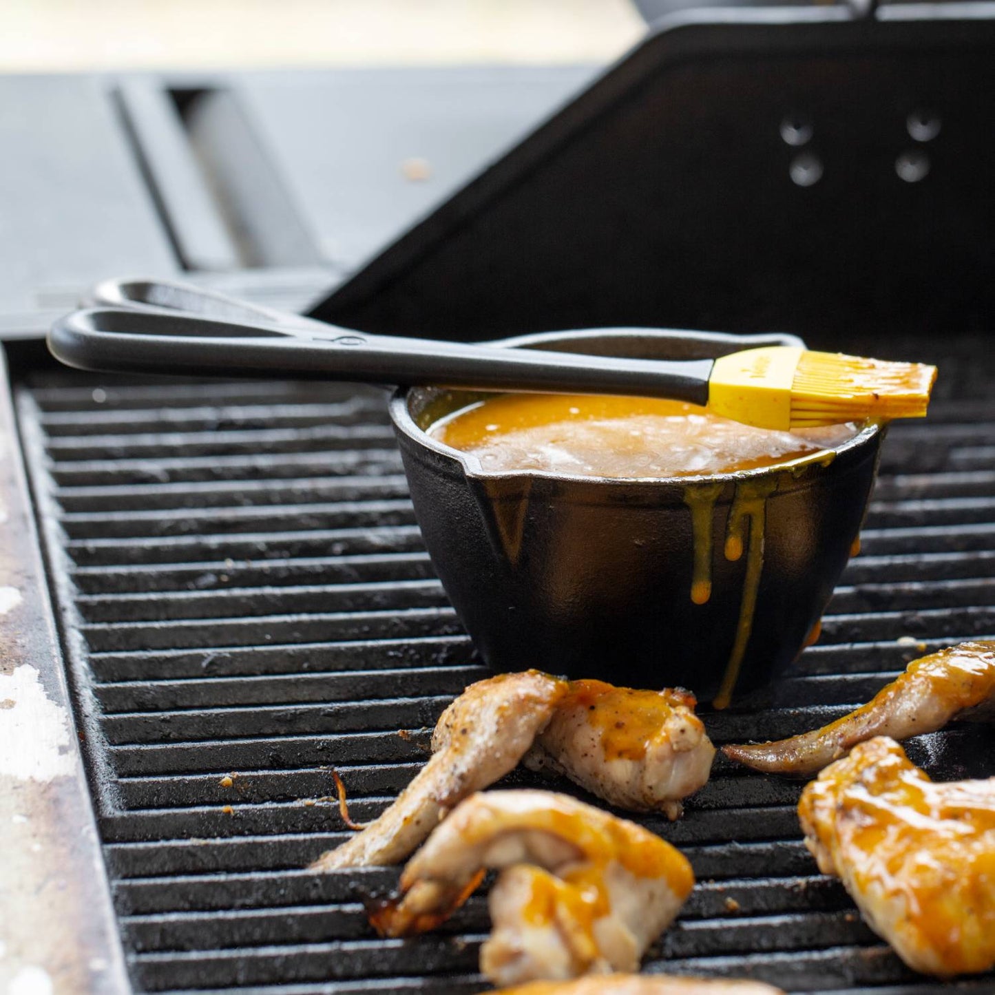 Black cast iron melting pot filled with sauce and topped with a silicone basting brush, placed on an outdoor grill alongside grilled chicken wings, ideal for Kitchen & Food and Cast Iron cookware collections.