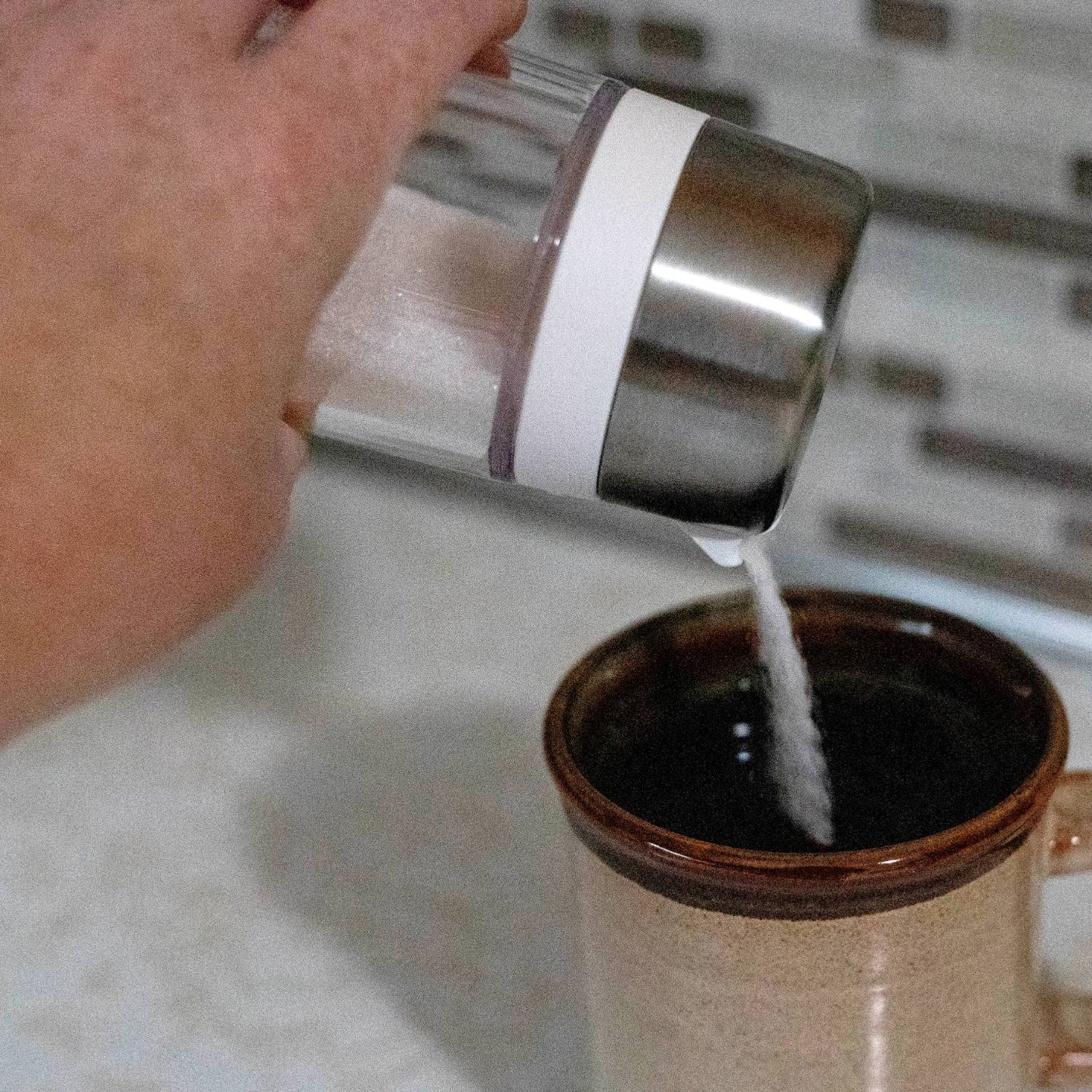 Hand pouring sugar from a clear glass dispenser with stainless steel lid and white band into a brown ceramic mug, designed to prevent clumping, ideal for Kitchen & Food and Kitchen Gadgets collections.