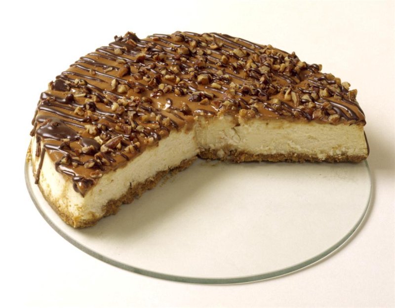 Slice of cheesecake topped with chocolate drizzle and chopped nuts on a clear glass plate, showcased for Kitchen & Food, Cookware, Baking Supplies, Best Sellers, Promo Eligible collections.