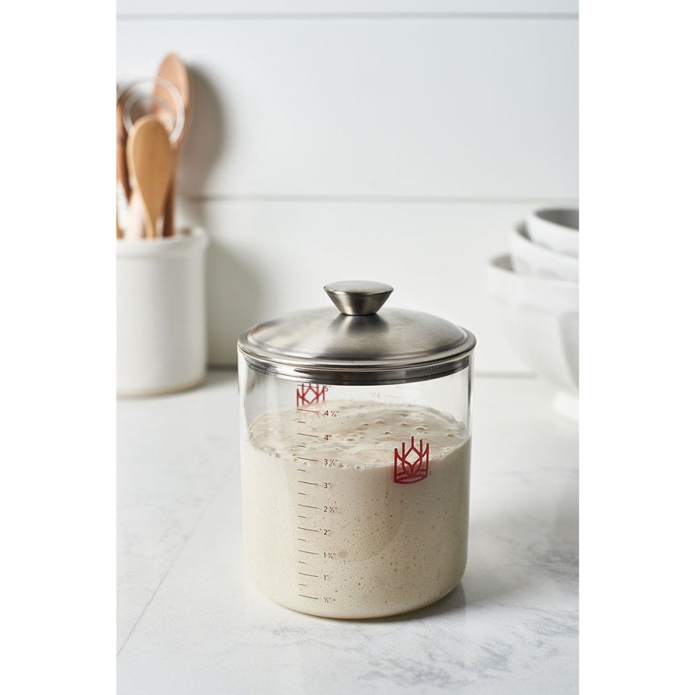 Clear glass sourdough crock with stainless steel lid filled with active starter, featuring red measurement markings, set on a white kitchen countertop with wooden utensils and stacked bowls nearby, suitable for Kitchen & Food, Cookware, Baking Supplies, Storage and Serving, and Best Sellers collections.