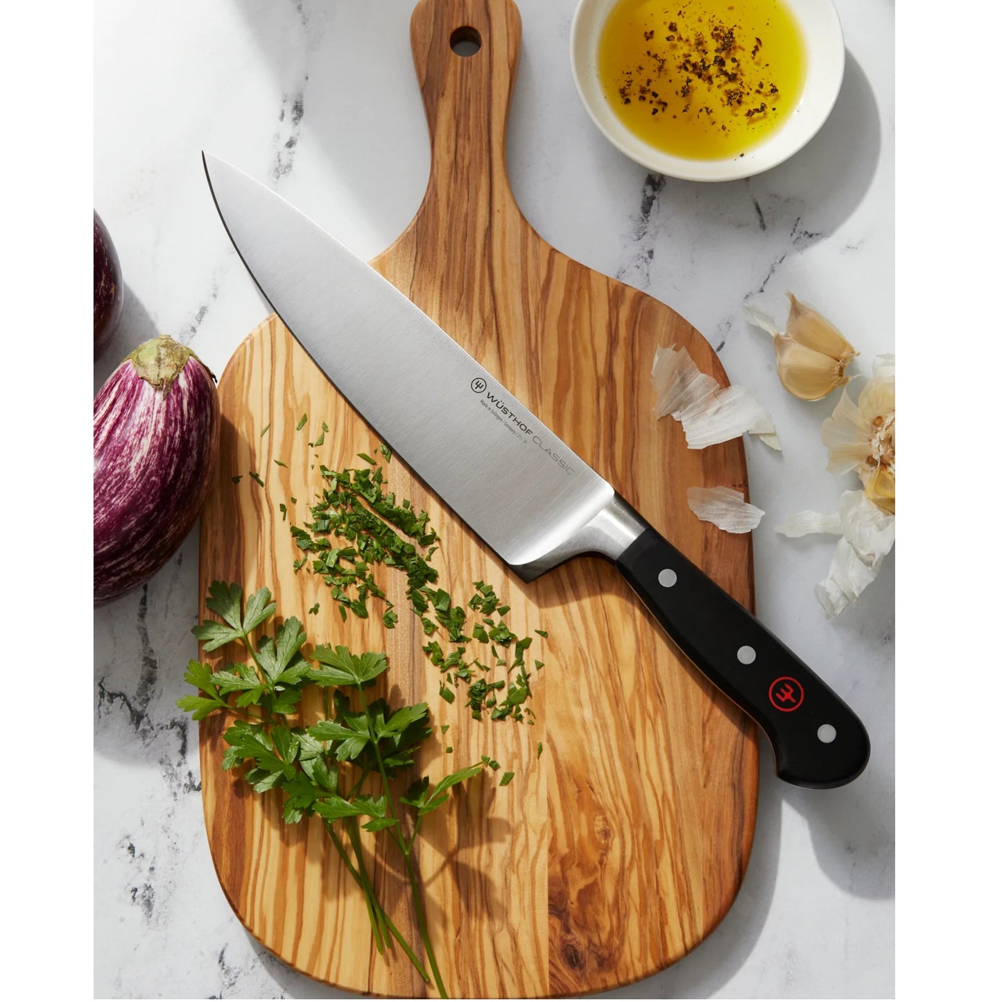 Sharp chef's knife with high carbon stainless steel blade and triple-riveted full tang handle resting on a wooden cutting board with chopped herbs, garlic, and olive oil, ideal for Kitchen Knives, Food Prep, and Best Sellers collections.