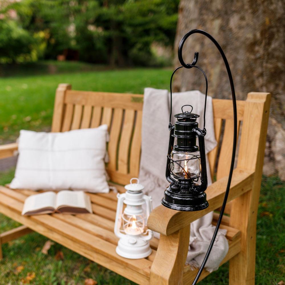 Black metal lantern holder with curved hook suspending a lit lantern outdoors on a wooden bench armrest, featuring a two-piece design for easy assembly, ideal for camping and emergency supplies.