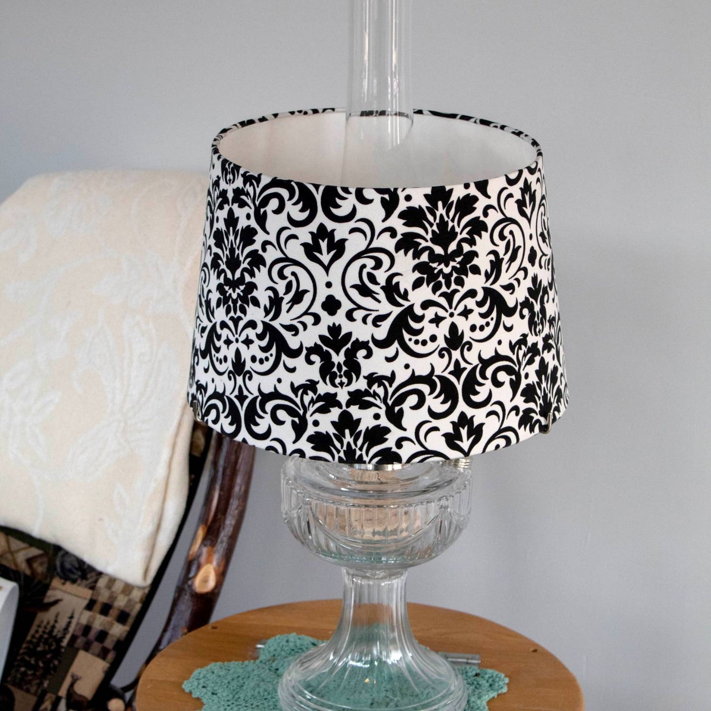 Black and white damask pattern cloth lamp shade designed for Aladdin oil lamps, shown on a clear glass lamp base in a cozy home setting, part of Lamps & Lighting collections.