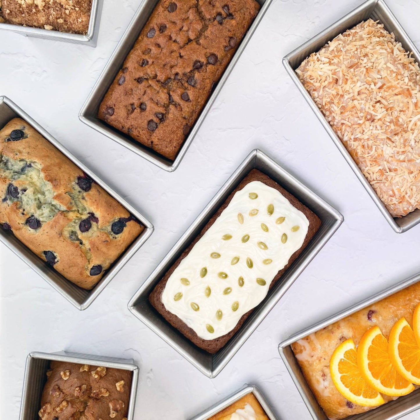 Collection of large loaf pans made from durable recycled steel, each filled with different baked goods including frosted, nut-topped, and fruit-adorned breads, ideal for Kitchen & Food and Baking Supplies collections.