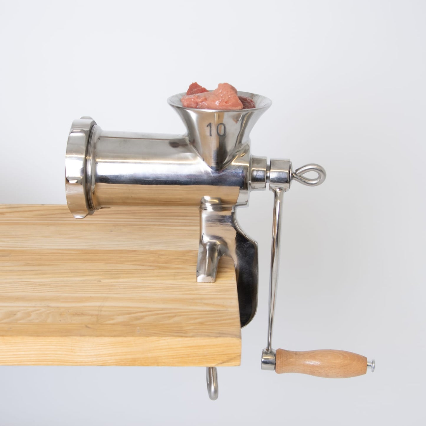 Stainless steel hand-cranked meat grinder with clamp attached to a wooden table, filled with fresh meat chunks, suitable for Kitchen & Food, Specialty Equipment, Butchering Supplies, and Best Sellers collections.