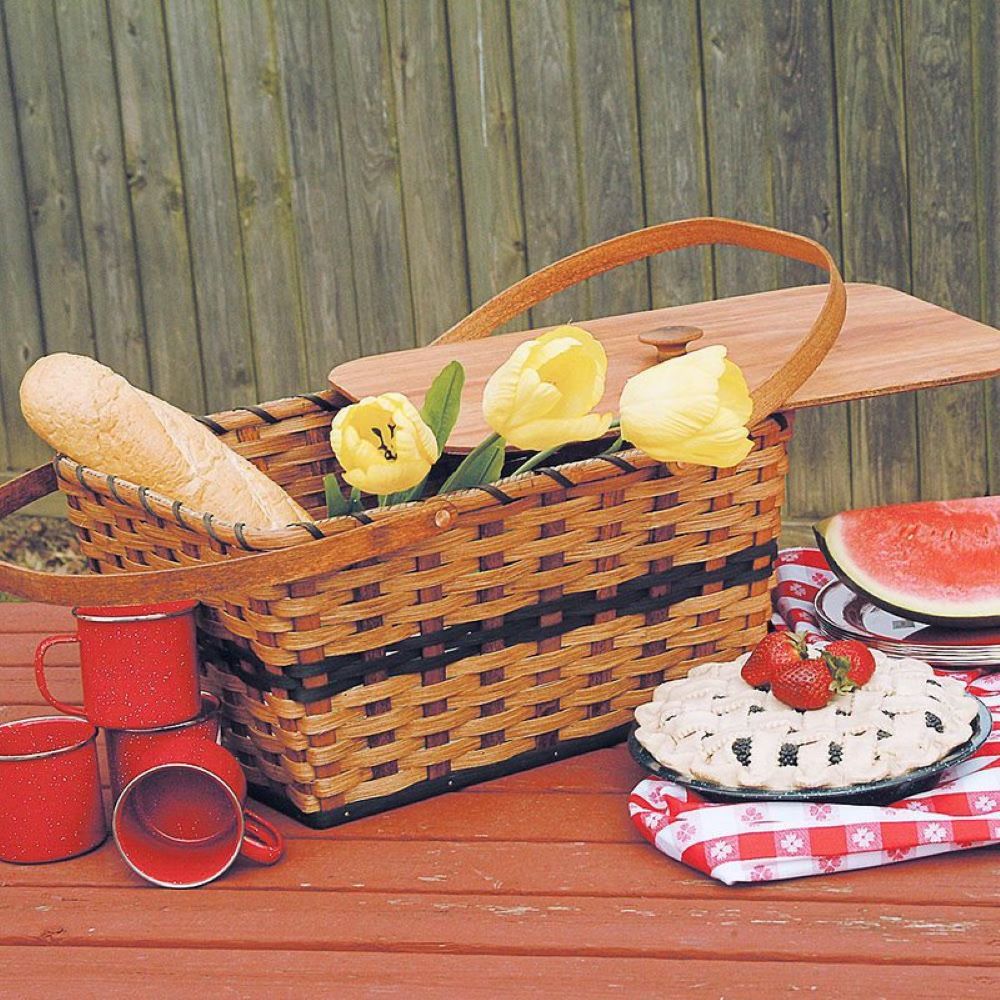 Handcrafted Amish woven basket with light-stained reeds and hunter green accents, featuring plywood lid and handles, filled with bread and yellow tulips, set for picnic in outdoor farm setting.
