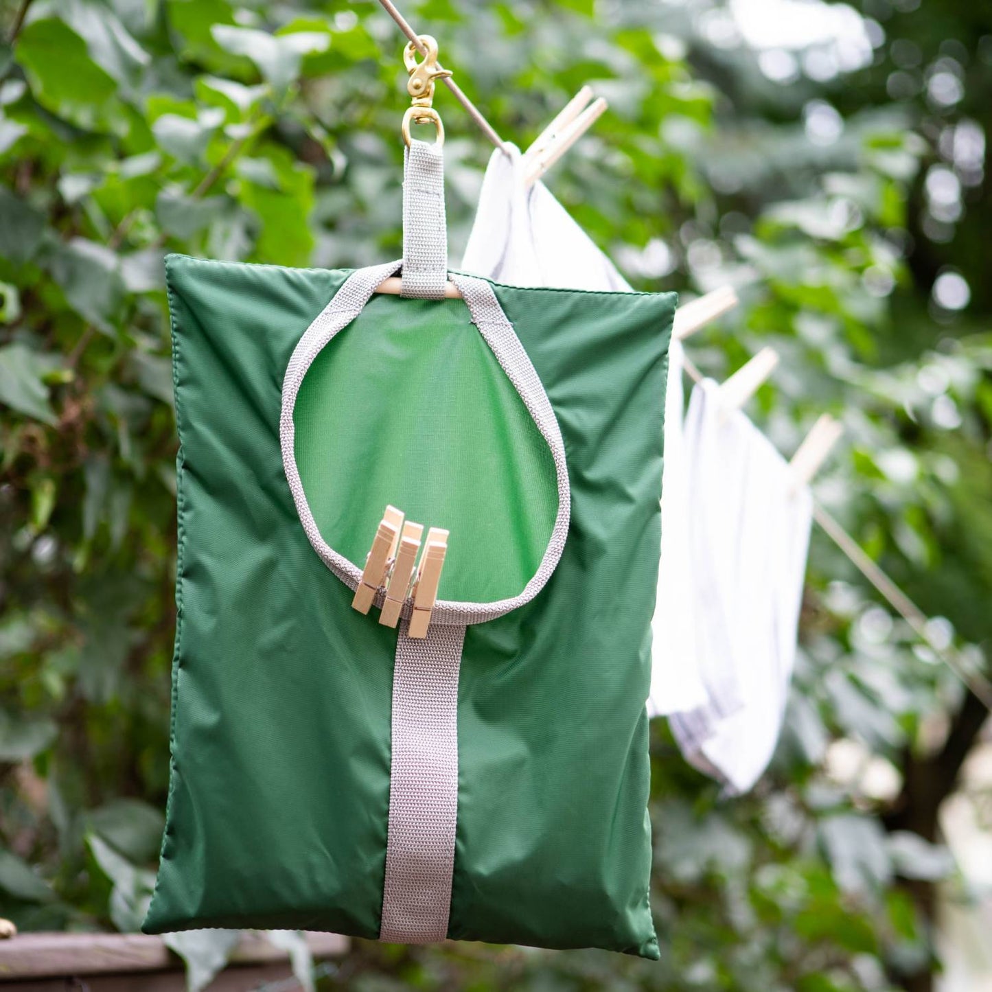 Green Amish clothespin bag with beige woven strap and brass clip hanging on clothesline, filled with wooden clothespins, handcrafted in USA for Home & Decor and Laundry Supplies collections.