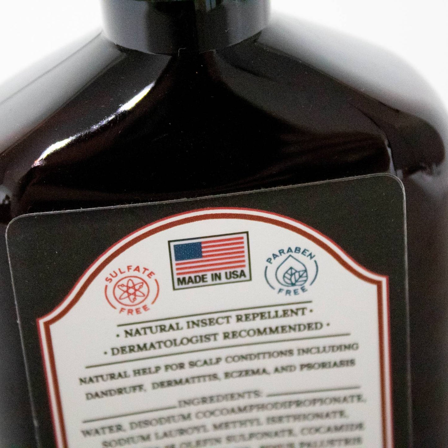 Close-up of a dark amber bottle label highlighting sulfate-free and paraben-free claims, made in USA, promoting natural insect repellent and dermatologist-recommended scalp care in Health & Wellness and Grooming collections.