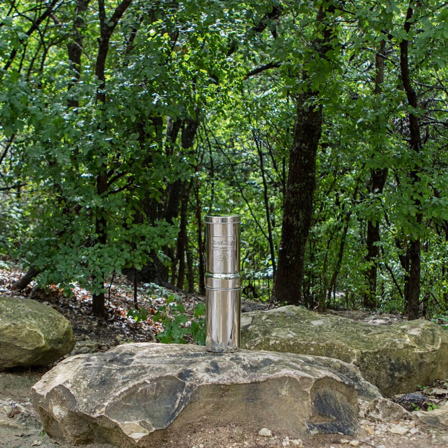 Polished stainless steel portable water filter kit standing on a large rock amid green forest, ideal for camping, emergency supplies, and USA Made outdoor gear collections.