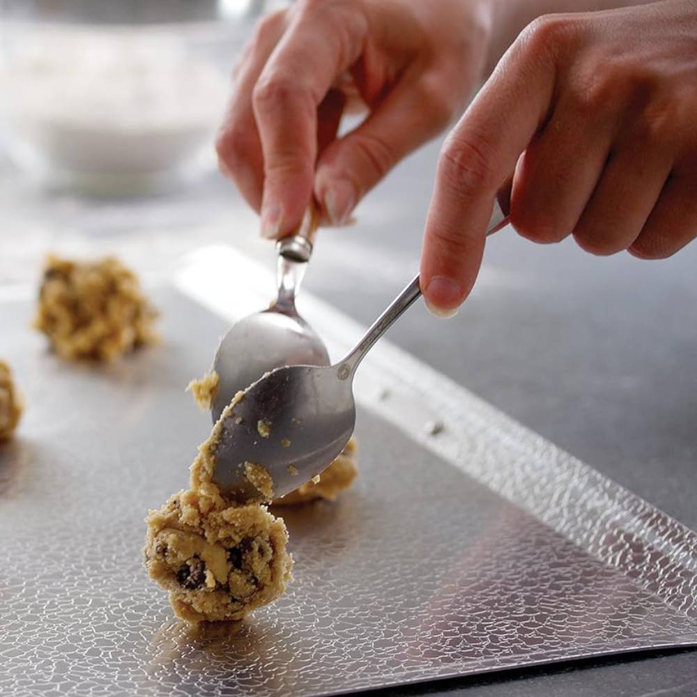 Human hands using two spoons to place cookie dough with chocolate chips onto a textured non-stick baking sheet, suitable for Kitchen & Food, Cookware, Baking Supplies, Pots and Pans, and Best Sellers collections.