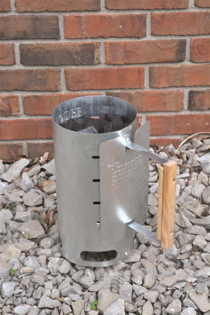 Aluminized steel charcoal starter with wooden handle standing on gravel against a brick wall, designed for quick briquet ignition, ideal for camping and farm tool collections.