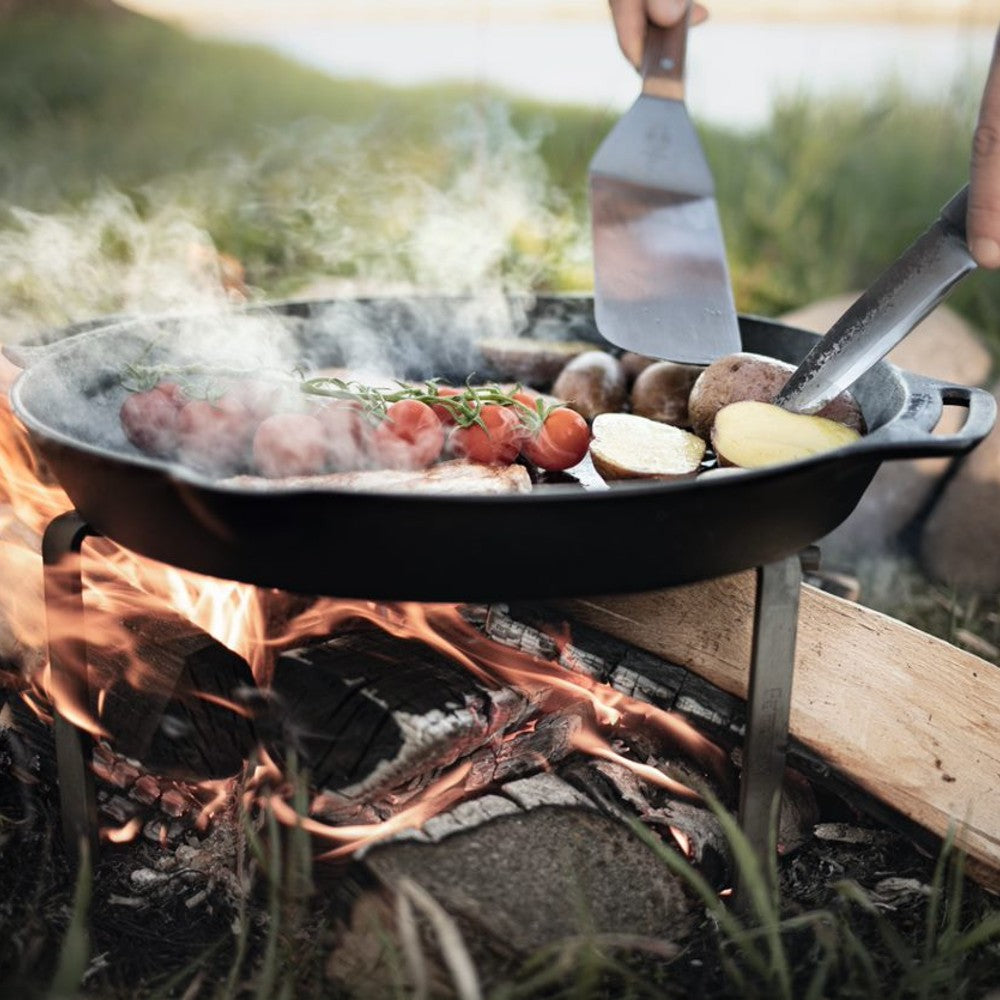 Heavy-duty steel cooking stand supporting a cast iron skillet over an open campfire with sausages, potatoes, and herbs, perfect for Farm, Garden & Tool, Camping, Best Sellers, and Promo Eligible collections.