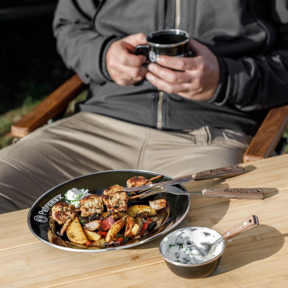 Black camping plate filled with grilled skewers and roasted vegetables, accompanied by wooden-handled fork and knife from a 5-piece camping cutlery set, alongside a small bowl of creamy dip on a wooden table in outdoor setting, part of Farm, Garden & Tool, Kitchen & Food, Camping, Dinnerware, and Best Sellers collections.