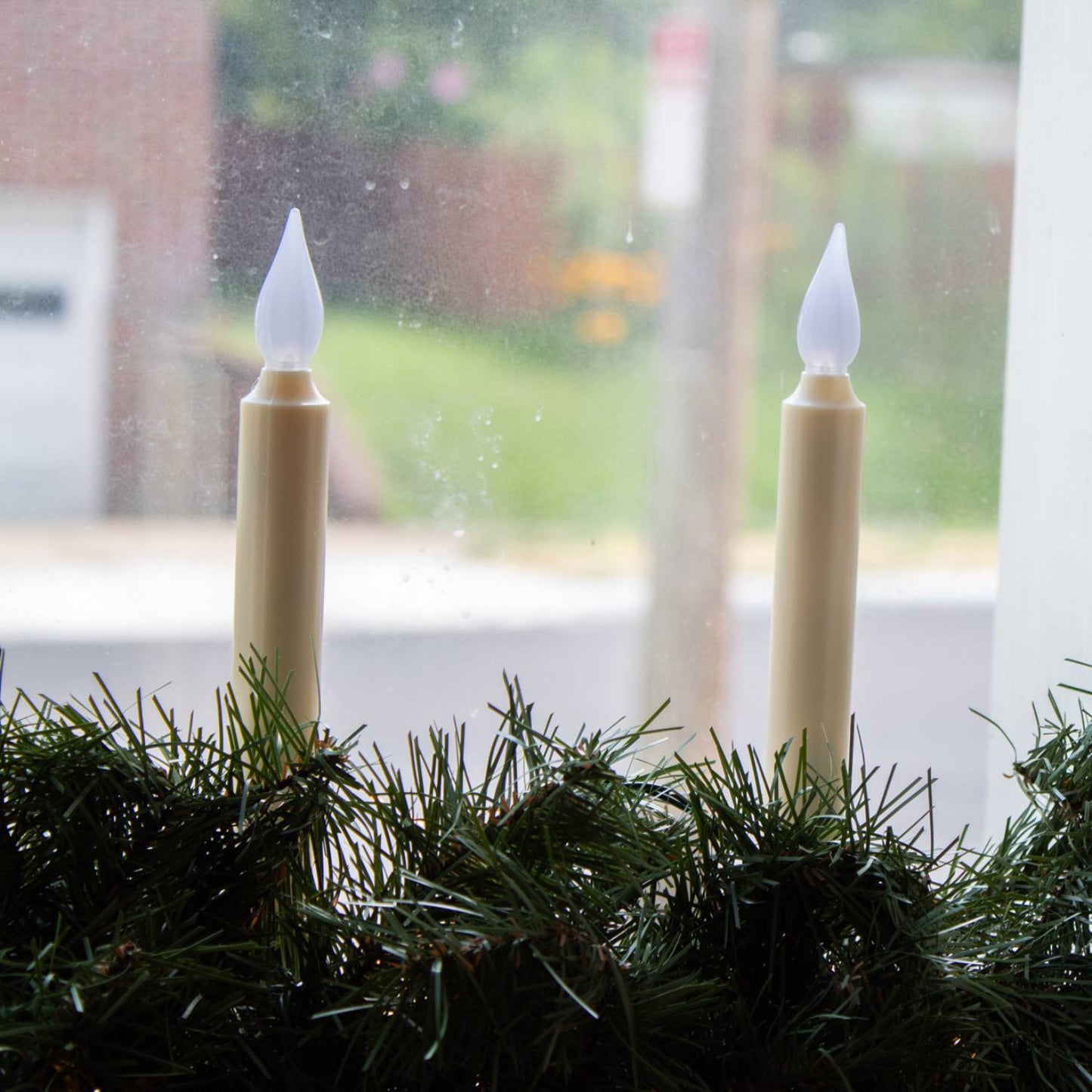 Pair of cordless LED taper candles with amber flickering flames atop slim bases nestled in green garland on a windowsill, perfect for holiday decor and best sellers collections.