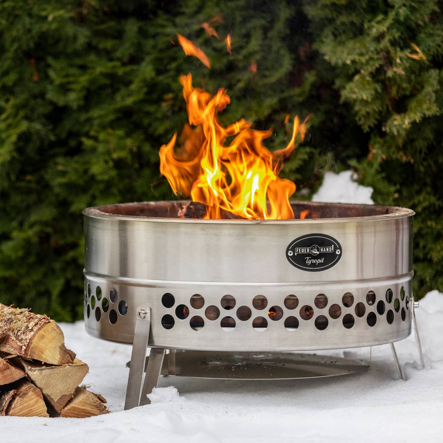 Stainless steel fire bowl with circular vent openings and sturdy legs, producing a lively flame outdoors on snow, ideal for camping and garden use in various collections.