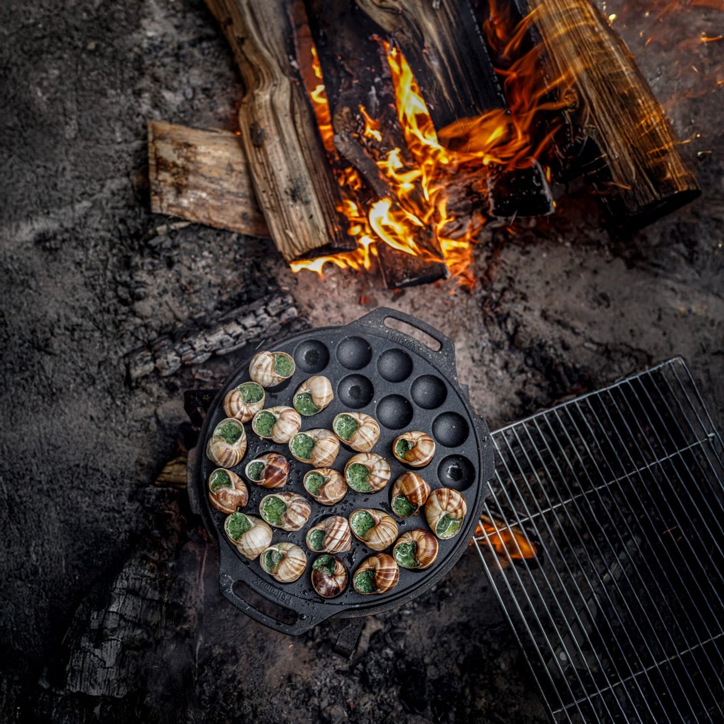 Cast iron poffertjes pan with 30 rounded molds cooking mini pancakes over an open fire, featuring two handles for safe outdoor use, ideal for camping, kitchen, and cookware collections.
