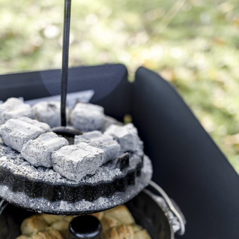 Black metal lifter with curved handle safely lifting a hot Dutch oven lid surrounded by glowing charcoal briquettes, ideal for Farm, Garden & Tool, Camping, and Best Sellers collections.