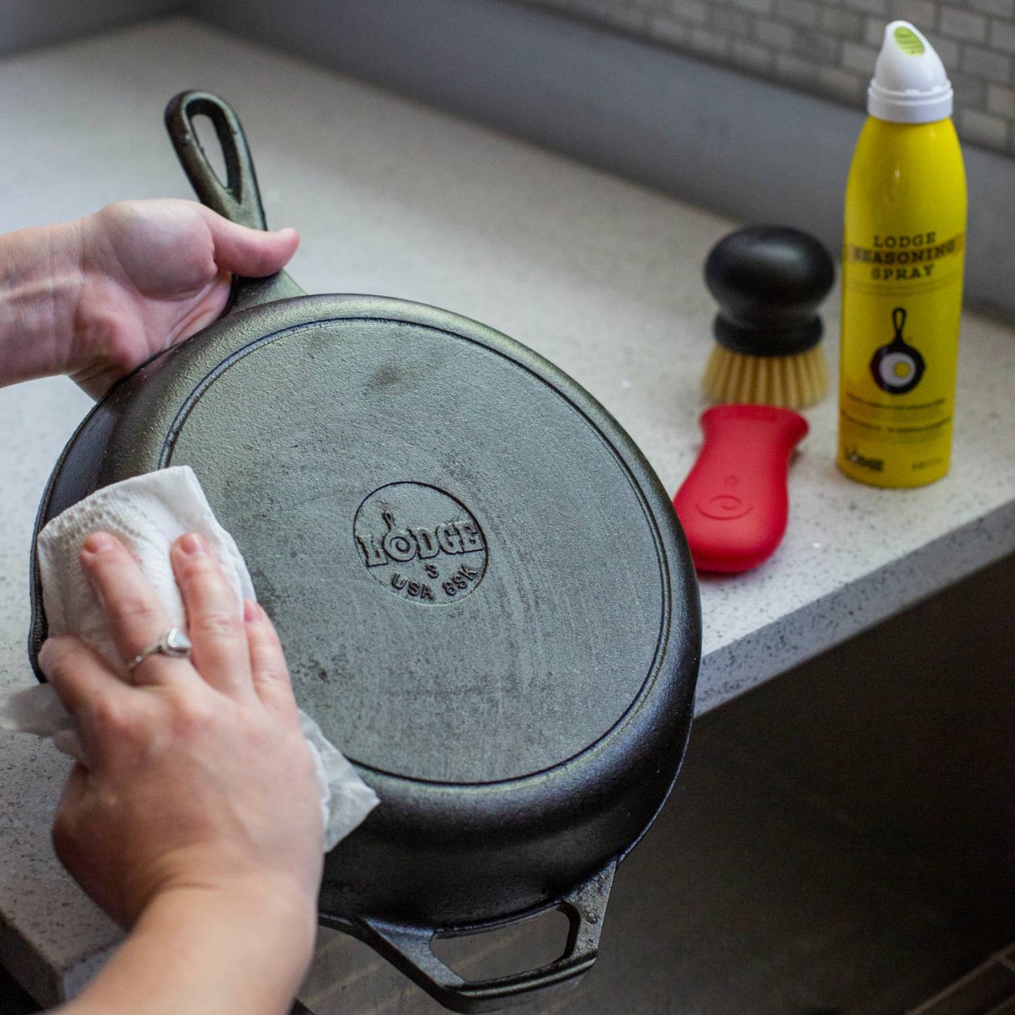 Person wiping the bottom of a Lodge cast iron skillet on a kitchen counter, with a yellow seasoning spray, a red handle silicone holder, and a nylon scrub brush nearby, ideal for Home & Decor, Kitchen & Food, and Cast Iron collections.