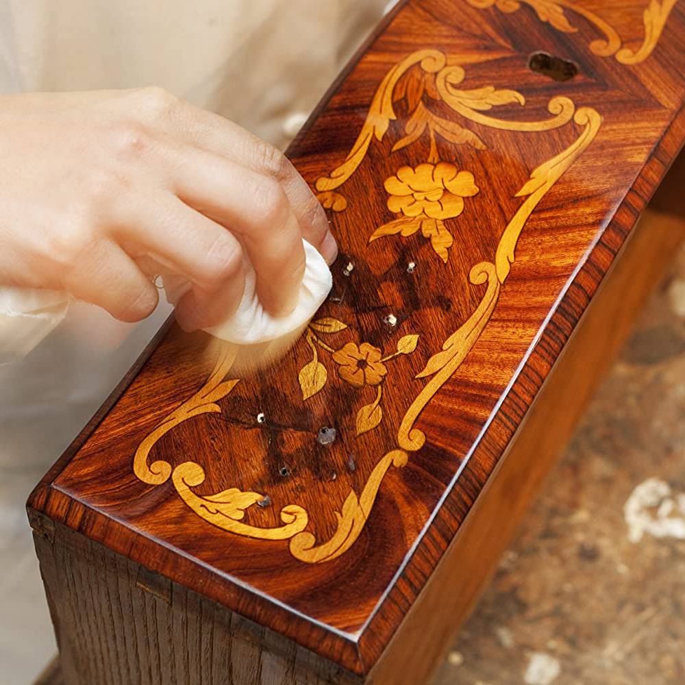 Hand applying handcrafted orange oil wood polish to a detailed wood surface with floral inlay, conditioning and enhancing the natural finish, ideal for Home & Decor and USA Made collections.