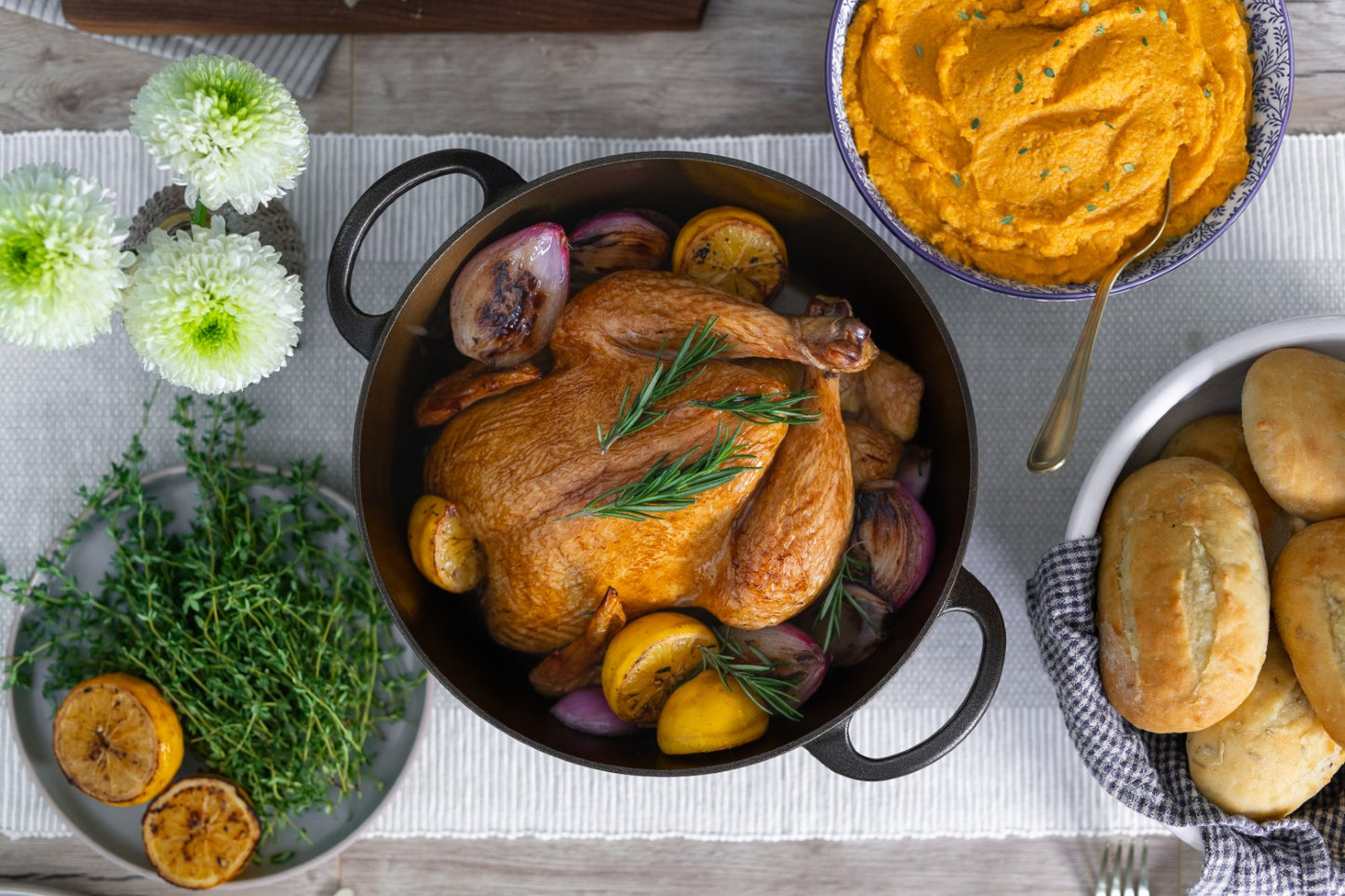 Handcrafted cast iron Dutch oven with roasted whole chicken, lemon halves, and herbs, surrounded by fresh thyme, green flowers, mashed sweet potatoes, and freshly baked rolls for Kitchen & Food, Camping, and USA Made collections.