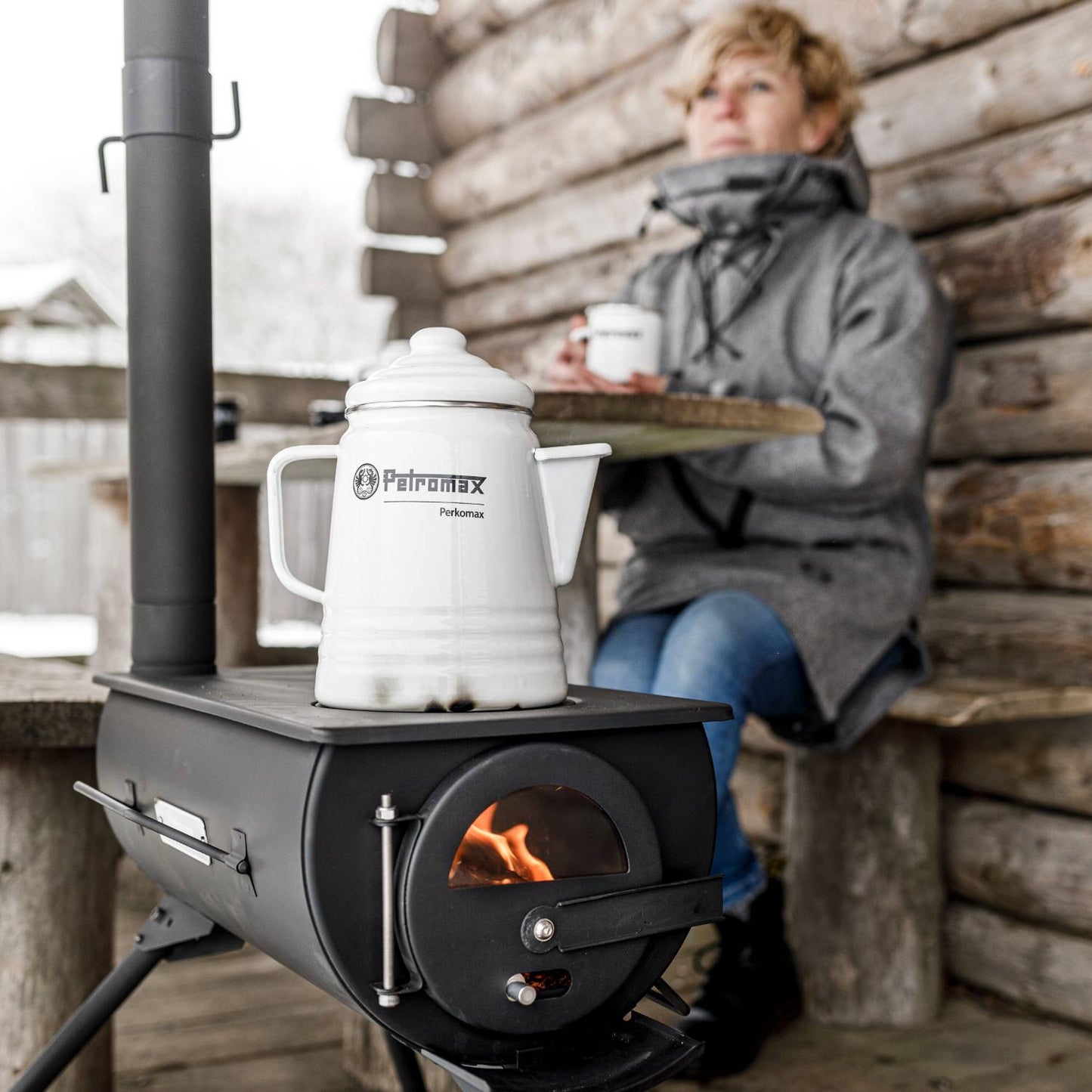 Black metal tent stove with visible fire burning inside, supporting a white enamel kettle, shown in rustic outdoor cabin setting with a person enjoying a warm drink, ideal for camping and farm use.