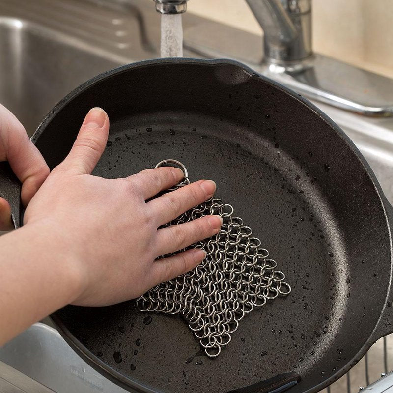 Hand using an extra large handcrafted stainless steel chainmail scrubber to clean a cast iron skillet under running water, part of kitchen cleaning and cookware accessories collections.