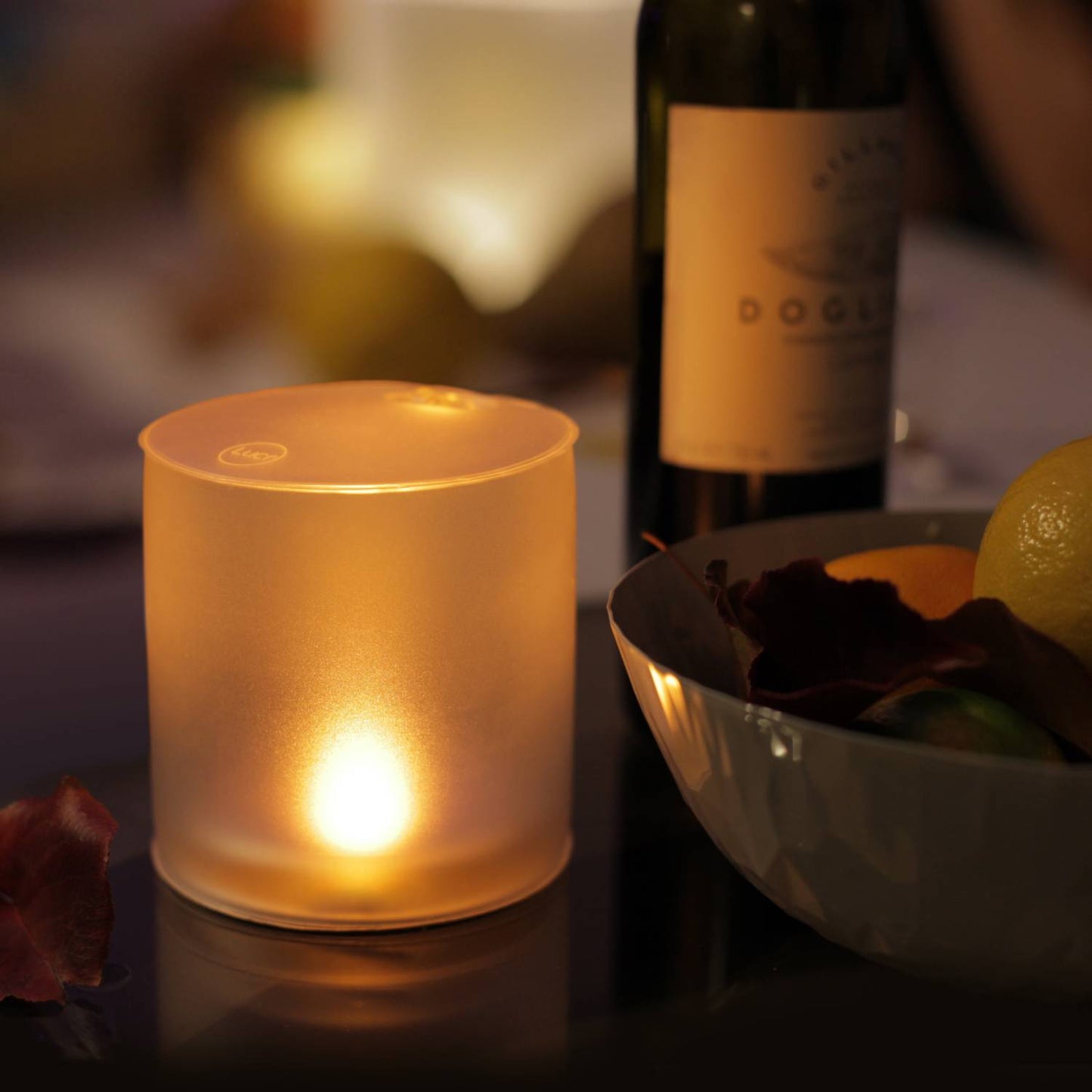 Soft warm light from a frosted inflatable solar LED candle flickering safely on a table next to a bottle and bowl, ideal for cozy outdoor or indoor ambiance in camping and emergency supplies.