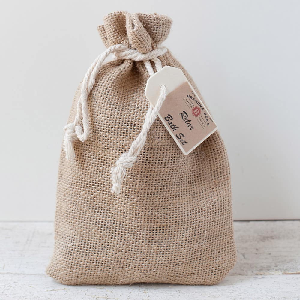 Natural burlap drawstring bag with a white rope tie and attached tag, containing a handcrafted USA-made relaxation bath gift set from Health & Wellness and Grooming collections.