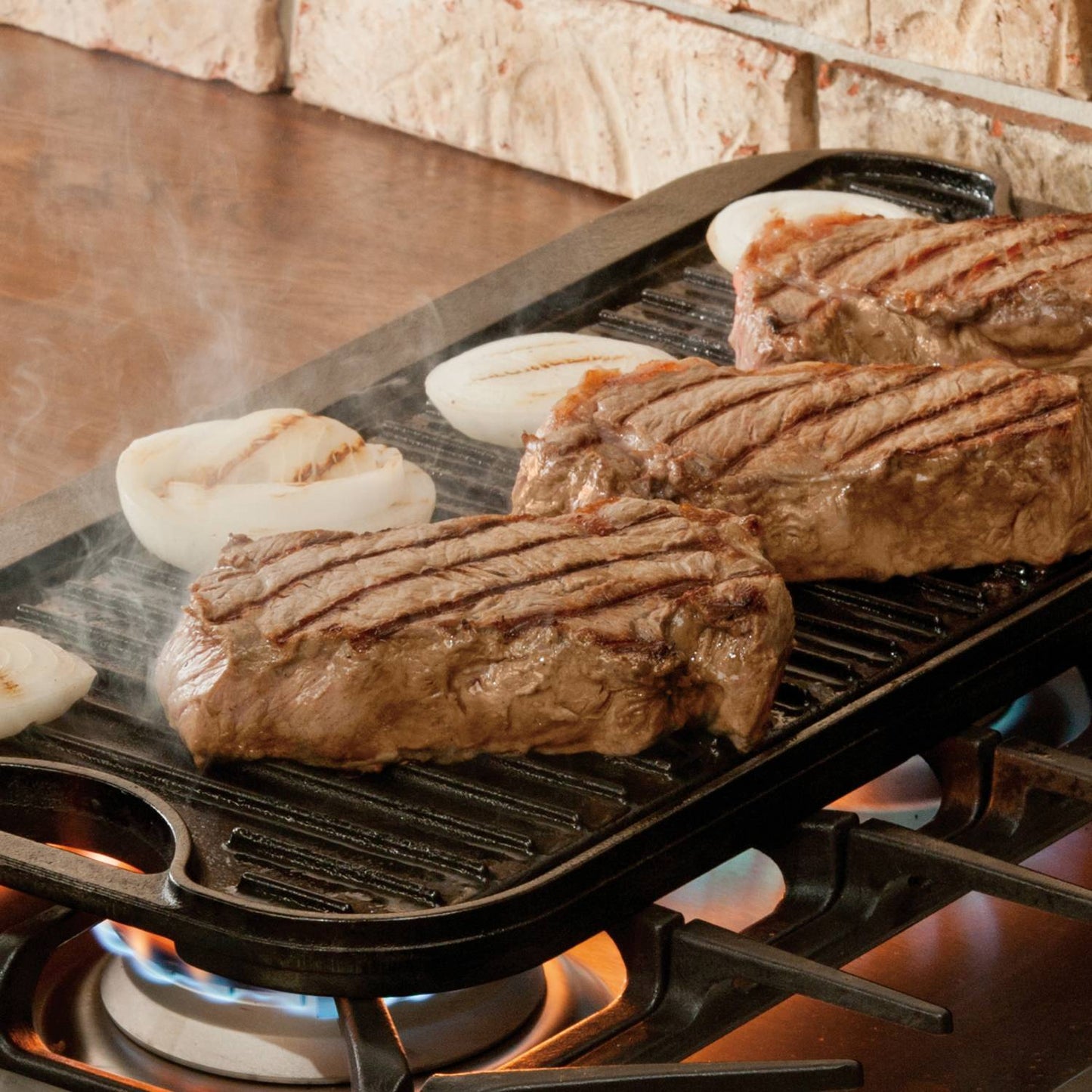 Cast iron dual-surface griddle with ribs and flat sides grilling seasoned steaks and onions over gas stove flames, featured in Kitchen & Food, Cast Iron, USA Made, and Best Sellers collections.