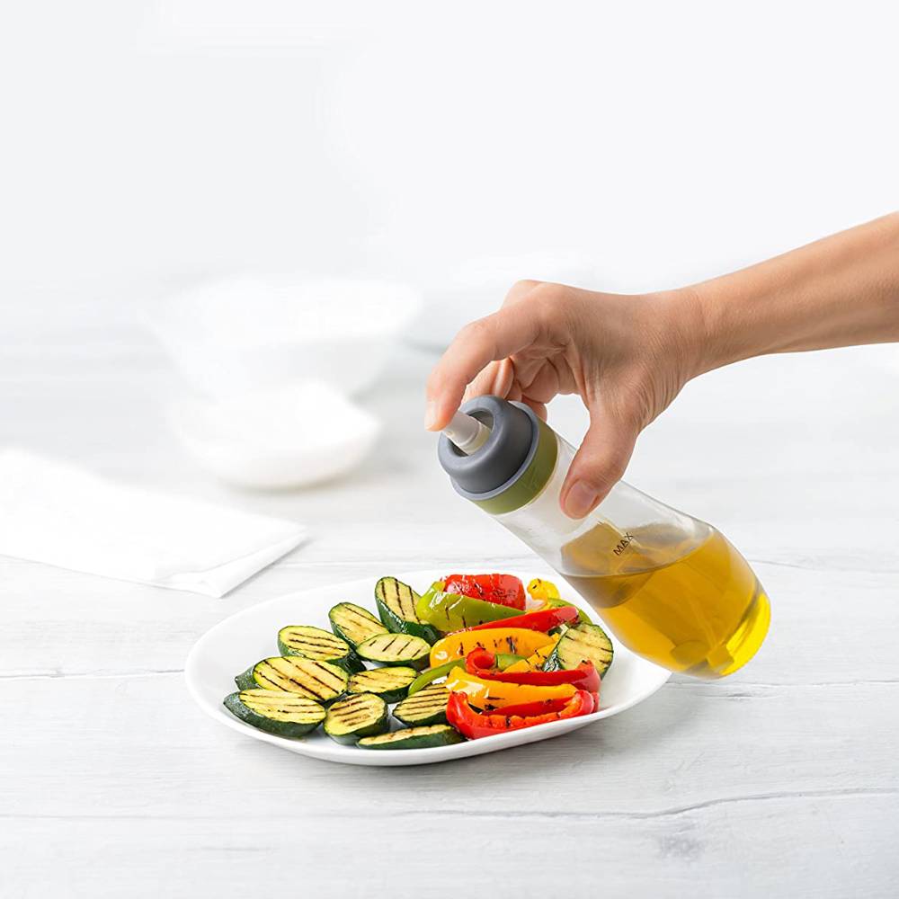 Hand dispensing oil from a clear glass spray bottle with a gray and green pump top onto grilled vegetables, suitable for Kitchen & Food, Cookware, and Baking Supplies collections.