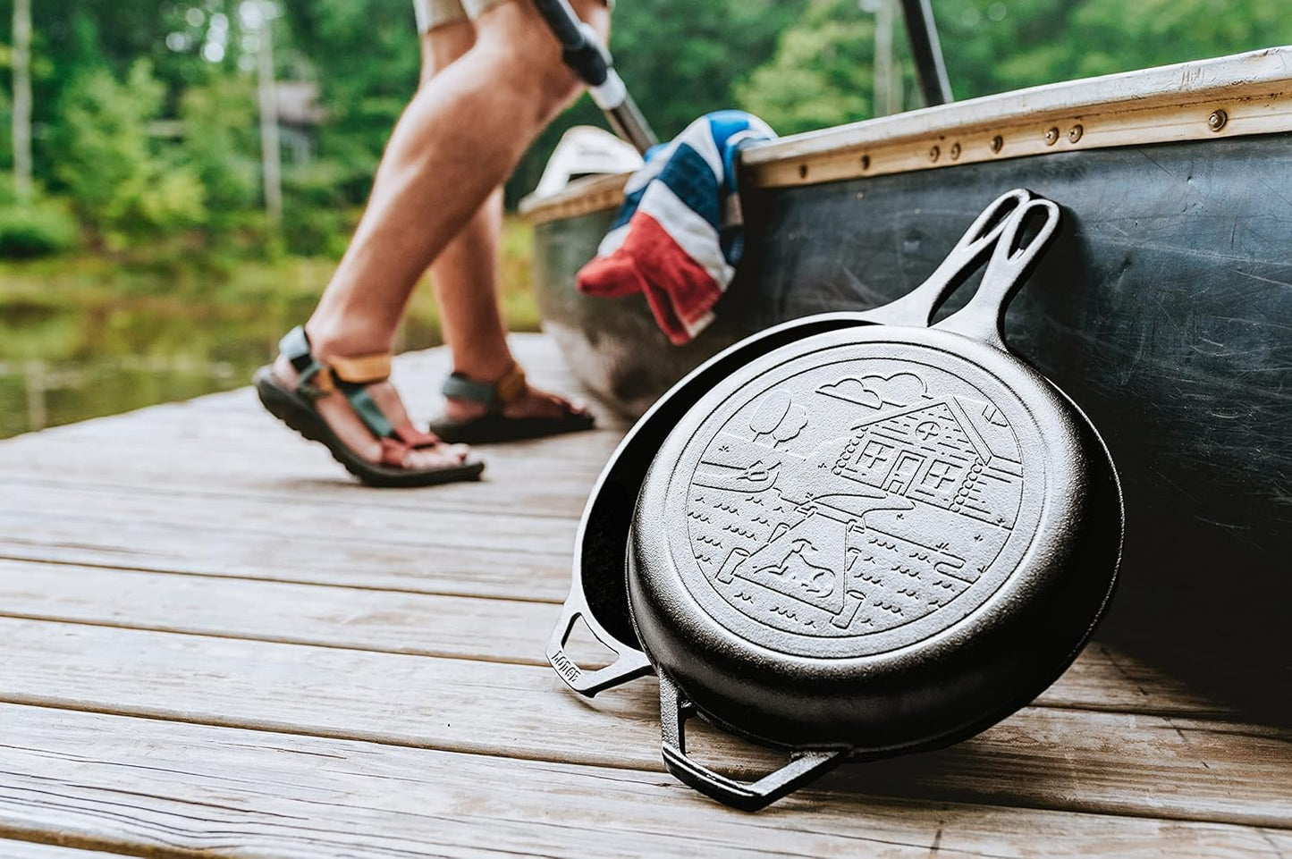 Seasoned cast iron combo cooker with an embossed cozy cabin design on the lid, resting on a wooden dock beside a canoe, perfect for camping, cookware, and outdoor cooking collections.