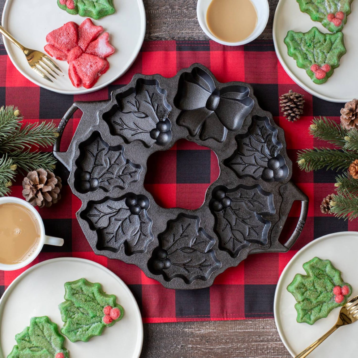 Seasoned cast iron cakelet pan featuring holiday wreath shapes with holly leaves and berries, resting on a red and black plaid cloth surrounded by festive baked treats and coffee, ideal for Kitchen & Food and Cast Iron collections.