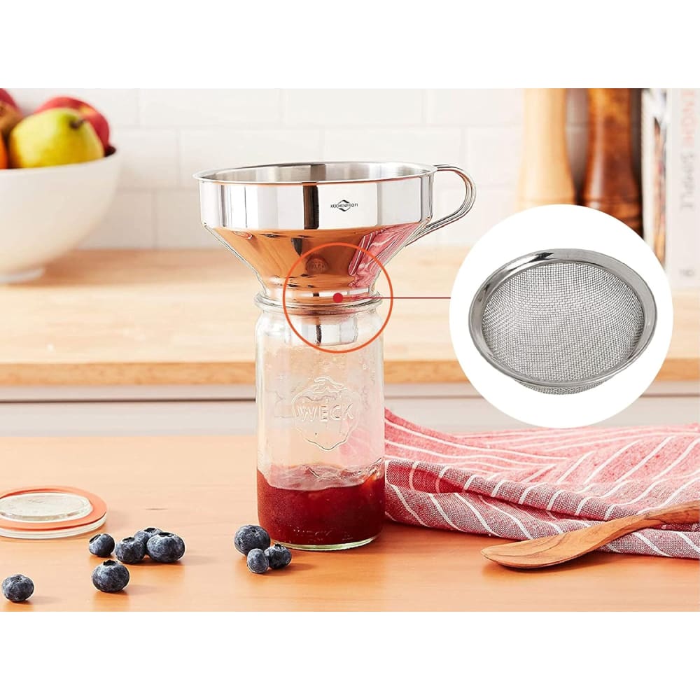 Stainless steel funnel with removable mesh filter set on a glass jar containing red liquid, surrounded by blueberries, wooden spoon, and kitchen towel in a bright kitchen setting.
