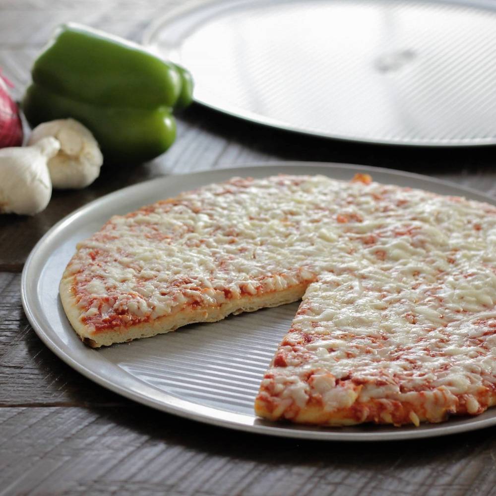 Crispy cheese pizza on a fluted aluminized steel pizza pan with a slice removed, surrounded by fresh garlic and green bell pepper, part of Kitchen & Food and USA Made collections.