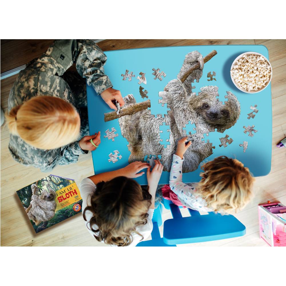 Three people working together on a uniquely shaped 100-piece sloth puzzle with large pieces on a blue table, alongside a book and a bowl of popcorn, ideal for children’s toys and games collections.