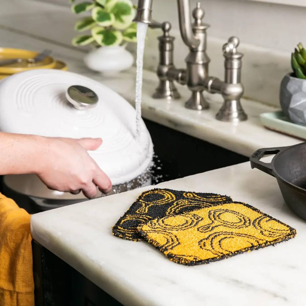 Two yellow and black scrub cloths with circular patterns, designed for gentle cleaning of cast iron and cookware, machine washable and made in USA, shown in a kitchen setting.