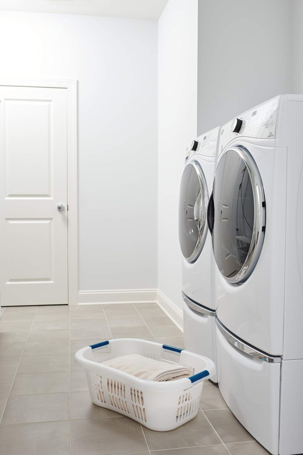 White plastic laundry basket with blue handles and ventilated sides, designed for easy carrying and hip support, placed on tiled floor beside front-loading washer and dryer in a minimalist laundry room.