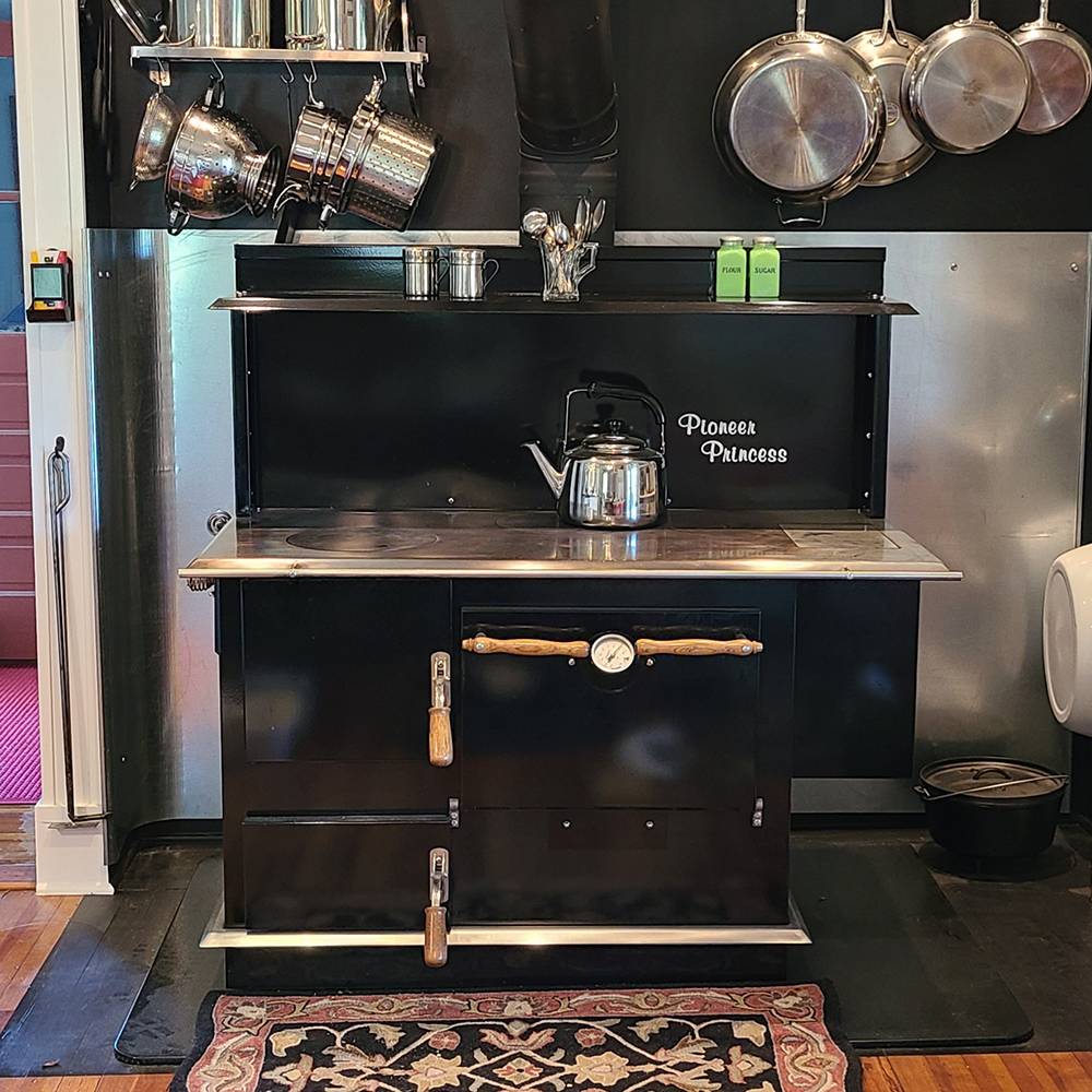 Black handcrafted wood cookstove with polished porcelain finish, wooden handles, large cooktop with a kettle, surrounded by hanging metal pots, perfect for Stoves, Cookstoves, and Best Sellers collections.