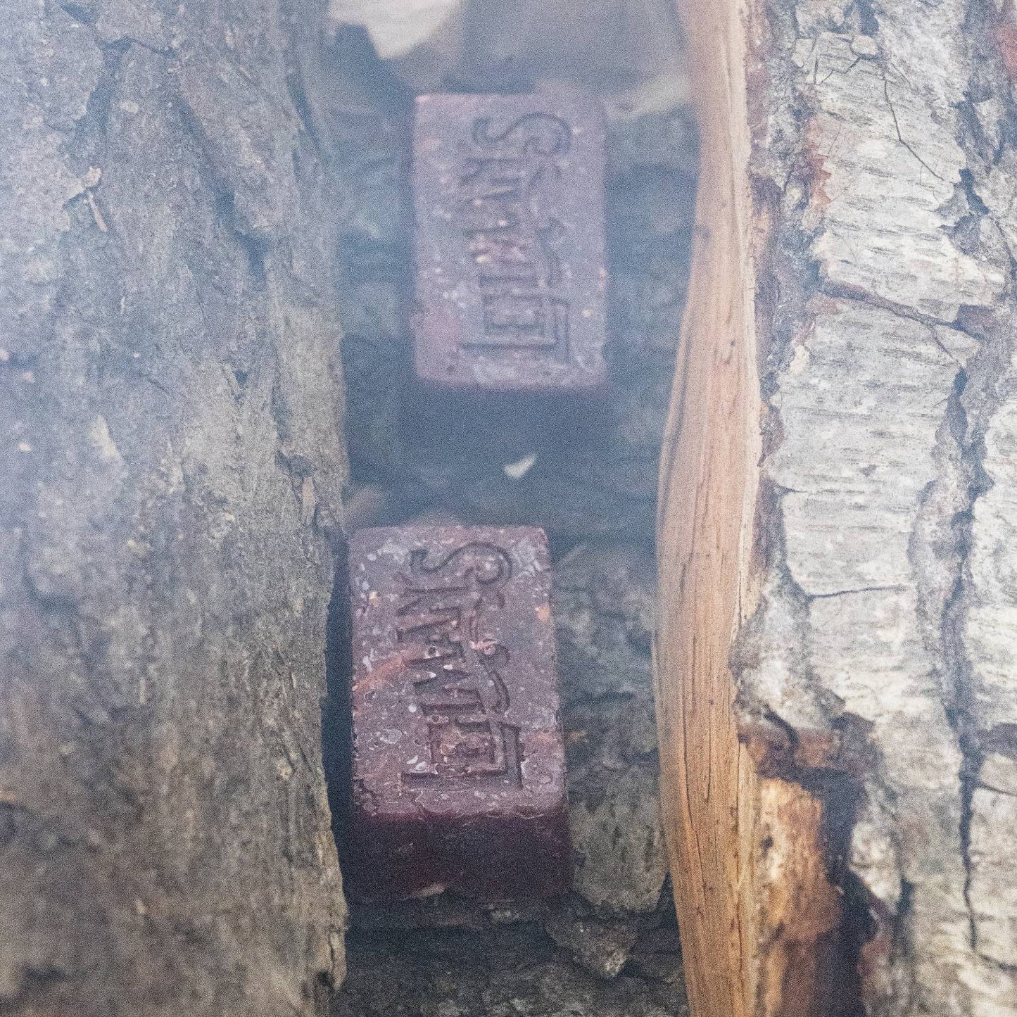 Two handcrafted reddish-brown fire starters embossed with Lehman's logo, nestled between split firewood, made in the USA for quick, non-toxic fire lighting in Camp, Hearth Accessories, and Stoves collections.