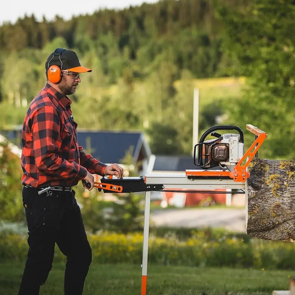 Man using an orange and black remote crank feed attachment to operate a gas chainsaw while cutting a thick log outdoors, suitable for Farm, Garden & Tool and Wood Cutting and Hauling collections.